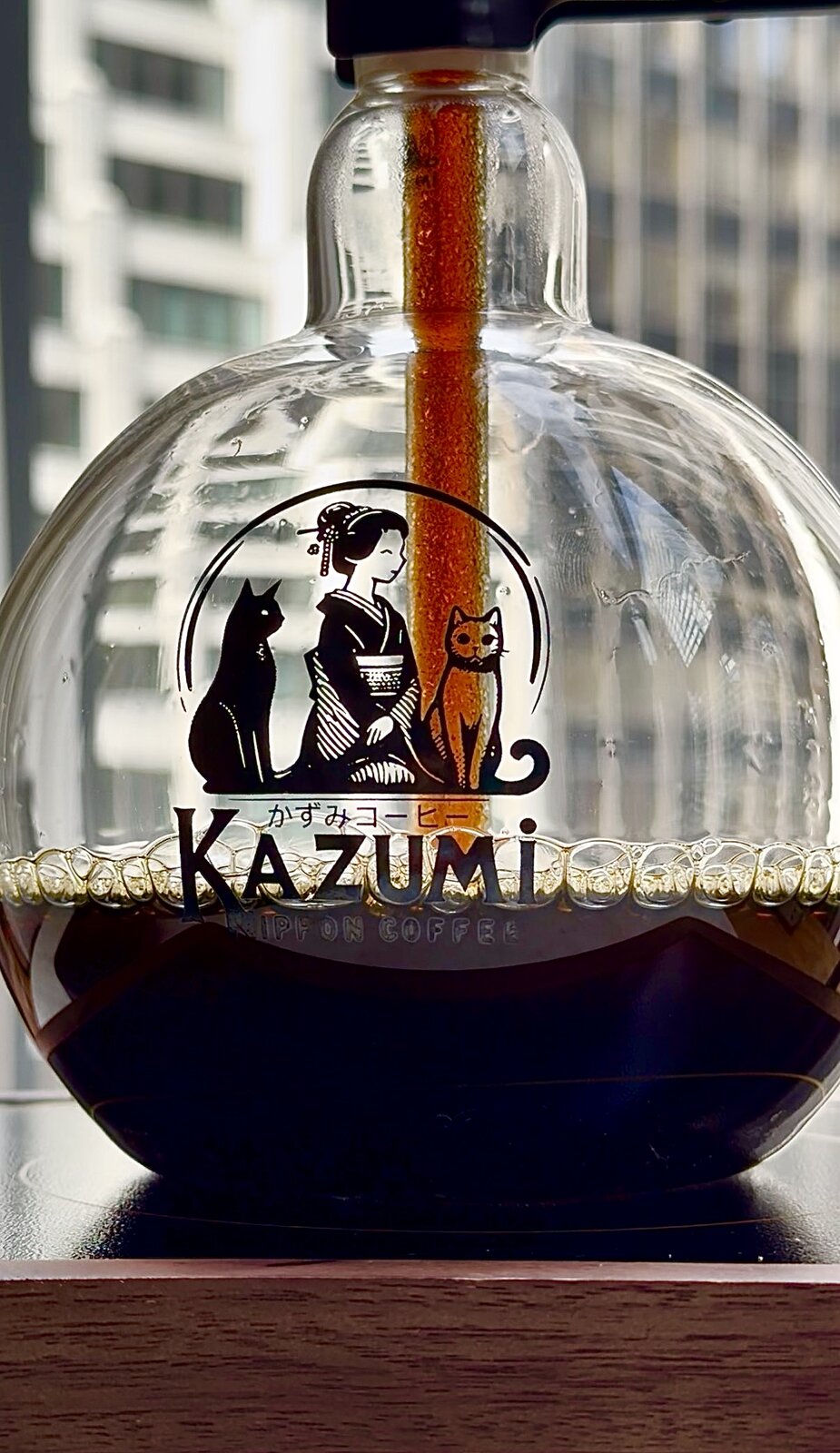 Kazumi all-glass siphon coffee maker brewing coffee against a New York skyline